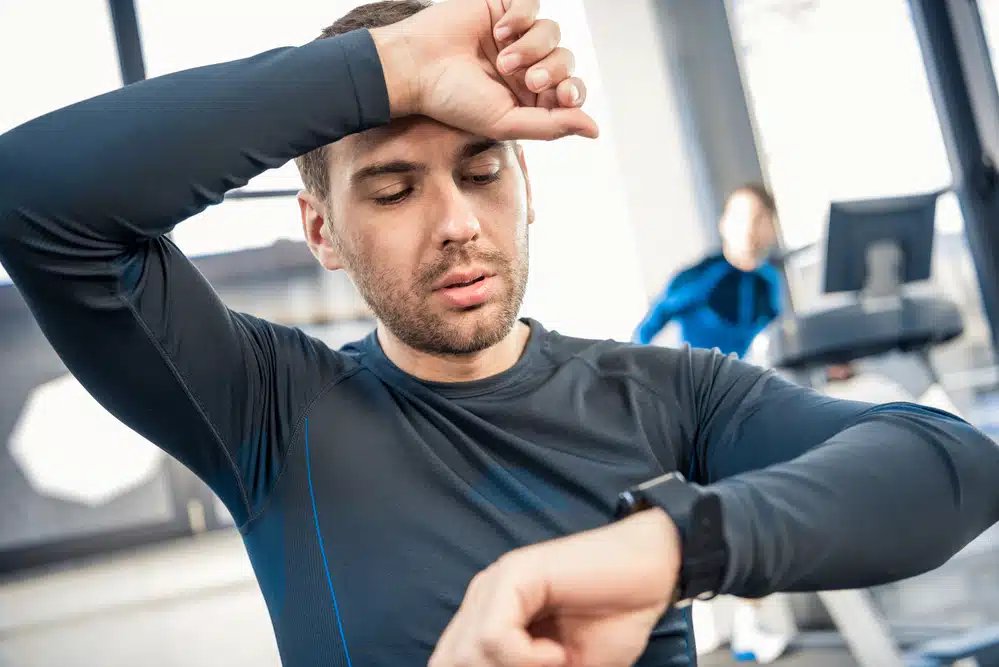 Un bărbat dintr-o sală de sport își șterge transpirația de pe frunte în timp ce își verifică trackerul de fitness, o altă persoană vizibilă în fundal pe o bandă de alergare.