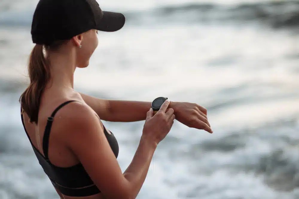 O femeie în sutien și șapcă sport negru își verifică ceasul smartwatch rezistent la apă pentru femei în timp ce stă lângă ocean.