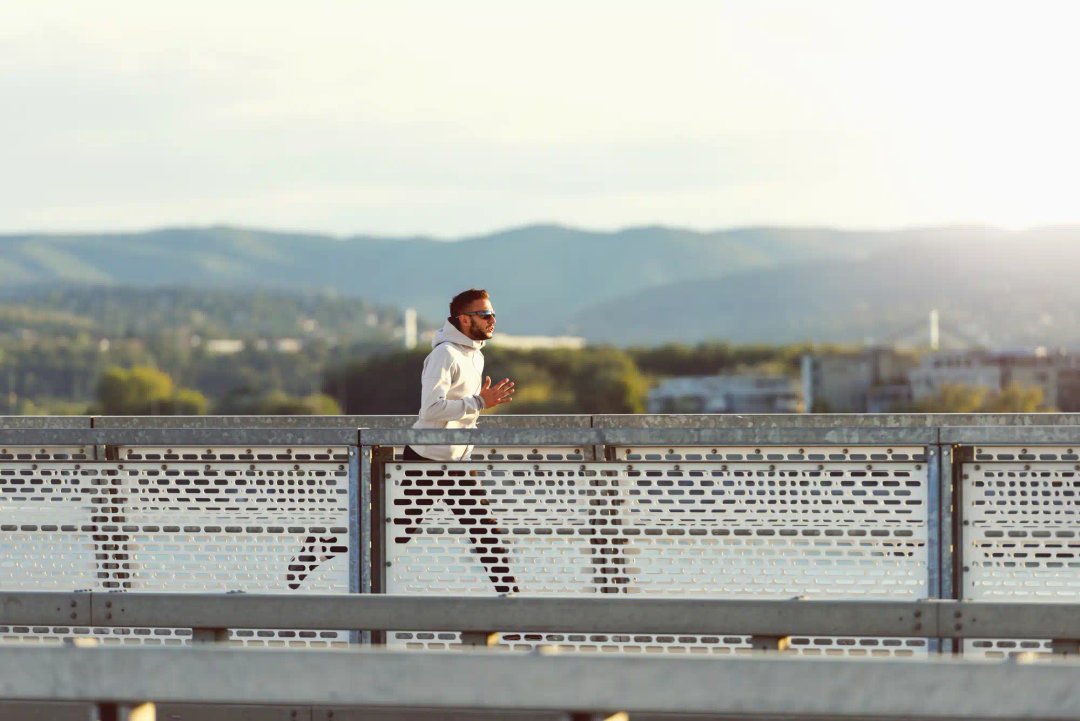Un bărbat face jogging pe un pod cu munți în fundal care poarta cele mai bune casti wireless bluetooth, casti alergare si casti sport.