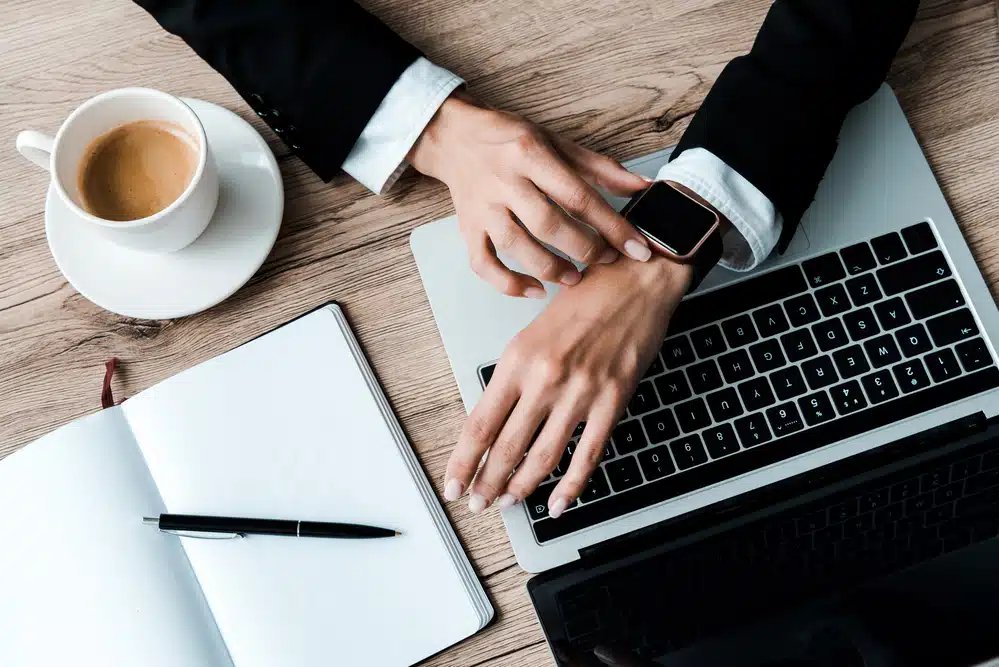 De ce nu se aprinde Smartwatch? O persoană care verifică un ceas inteligent în timp ce tastează pe un laptop, cu o ceașcă de cafea și un notebook pe un birou de lemn.