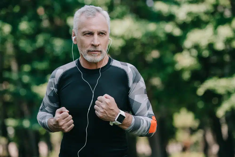 Bărbat în vârstă care face jogging într-un parc în timp ce ascultă muzică la căști, purtând o cămașă cu imprimeu camo și un ceas inteligent.