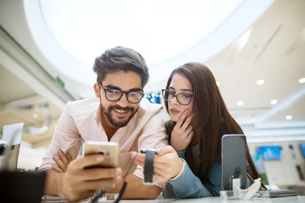 Un bărbat și o femeie fac cumpărături pentru ceasuri inteligente într-un magazin de electronice. Textul în limba română promovează calitatea, stilul și prețurile excelente, cu o notă despre opțiunile de livrare rapidă. Magazinul evidențiază, de asemenea, selecția lor de casti wireless pentru o experiență audio fără întreruperi.