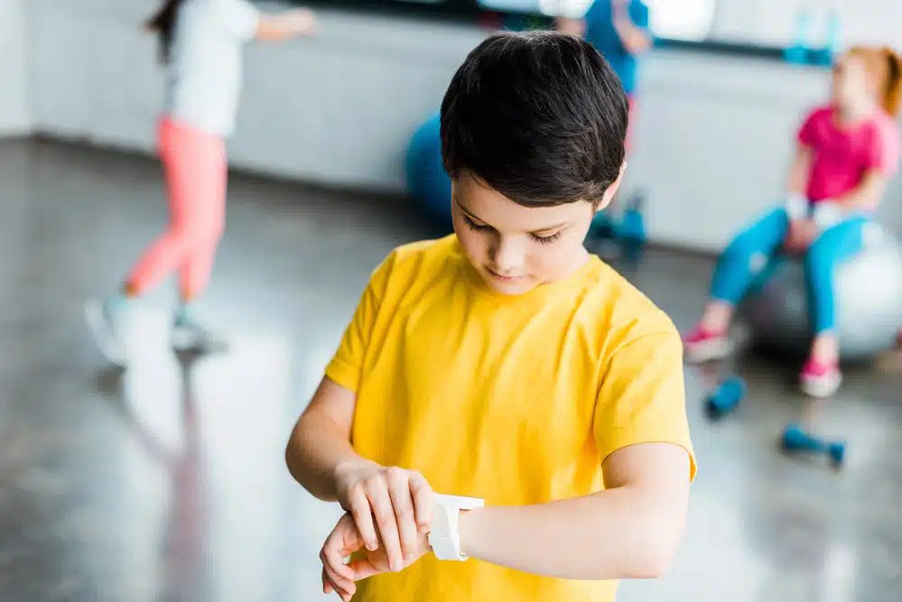 Un băiat într-un tricou galben își reglează un tracker de fitness pe încheietura mâinii într-o sală de sport, cu figuri neclare în fundal.