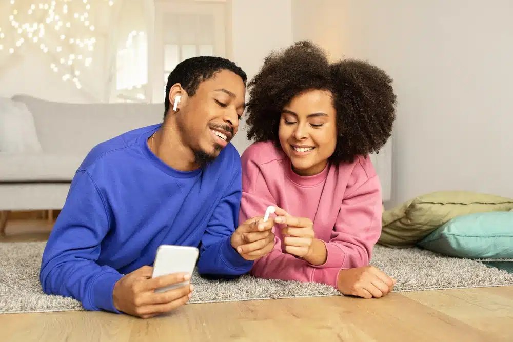 Un bărbat și o femeie stau pe un covor, zâmbind și împărțind o pereche de căști bluetooth conectate la un smartphone.