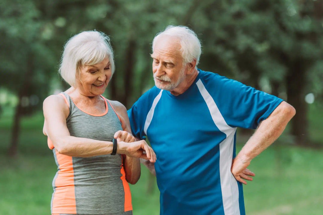O femeie în vârstă în îmbrăcăminte sportivă îi arată ceasul inteligent unui bărbat în vârstă într-o cămașă sport albastră în timp ce stă într-un parc.
